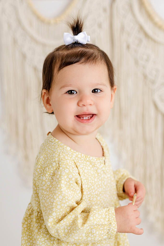 Smiling baby girl in yellow floral dress with white bow hair clip posing for a portrait photo.