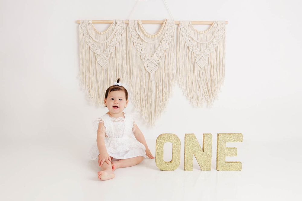Baby girl in white dress sitting next to gold glitter ONE letters for first birthday photo session.