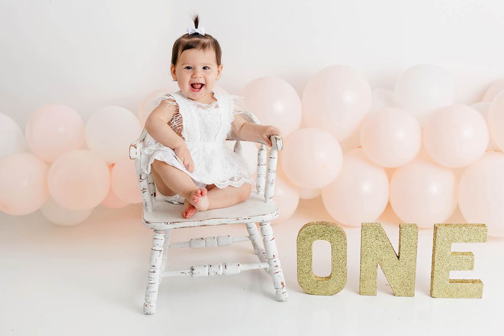 Baby girl in white dress sits on chair with pink balloons and glittery ONE sign for first birthday cake smash.