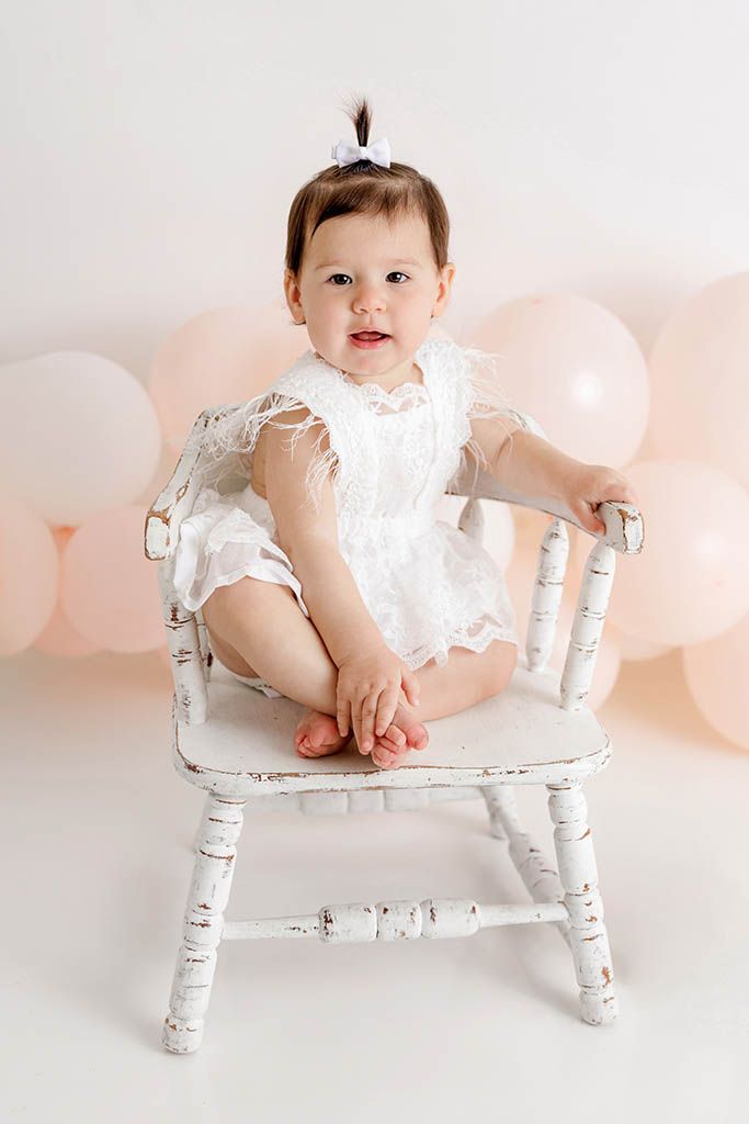 Baby girl in white lace romper sits on rustic white chair surrounded by pink balloons during first birthday photo session.