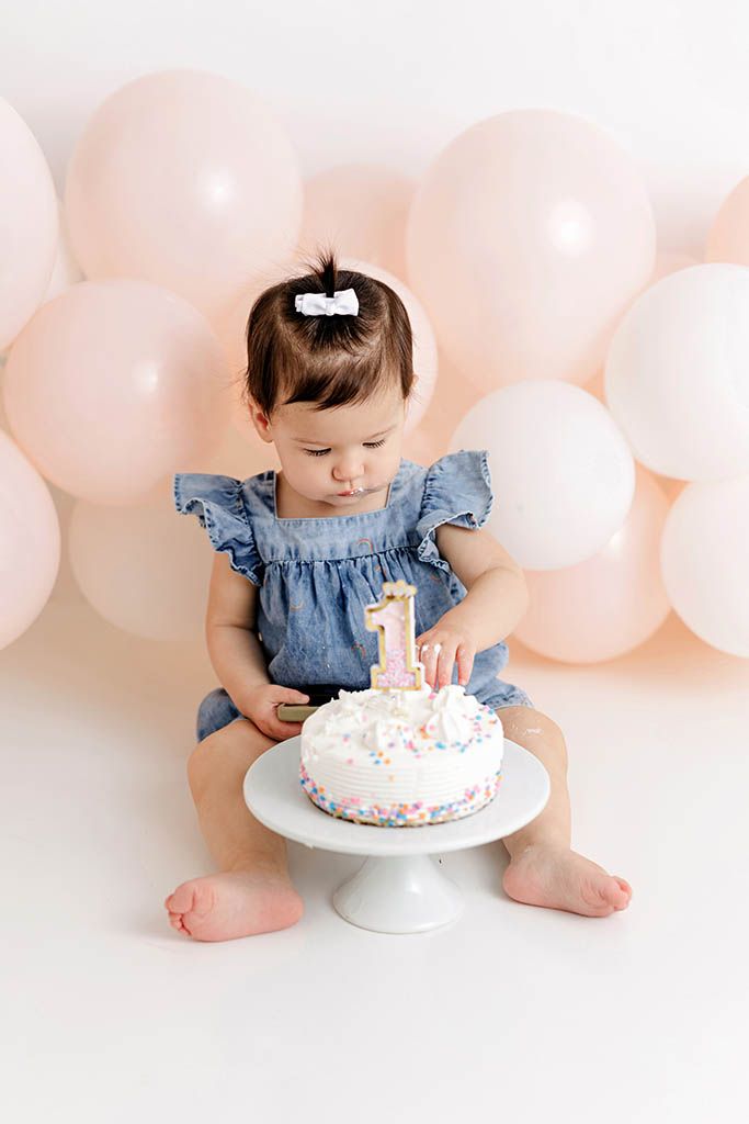 Baby girl in blue dress touching her first birthday cake with pink balloons backdrop.