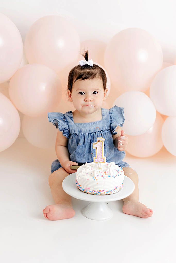 Baby girl in blue dress sits with first birthday cake and number 1 candle, surrounded by pink balloons.