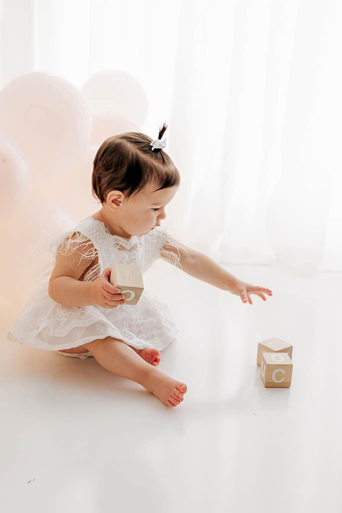 Adorable baby girl in white lace dress reaching for wooden alphabet blocks during birthday photoshoot.
