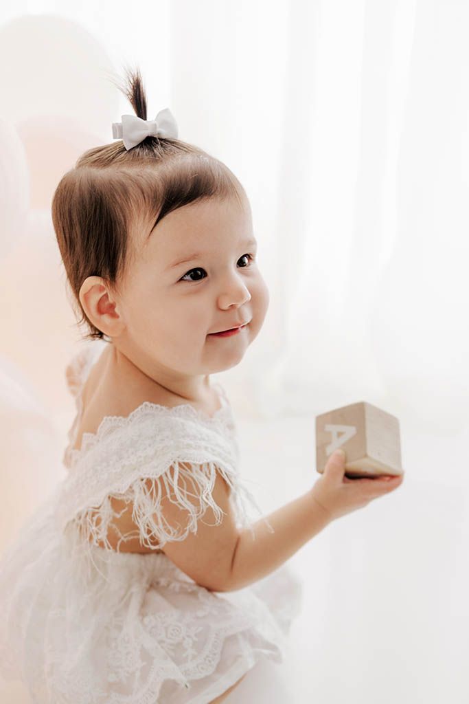 Cute baby girl in white lace dress holding a wooden alphabet block during a first birthday photoshoot.