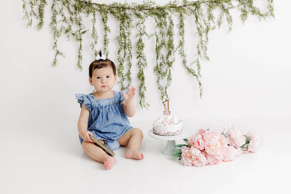 Baby girl in blue romper sits beside first birthday cake with pink peonies and greenery garland backdrop.