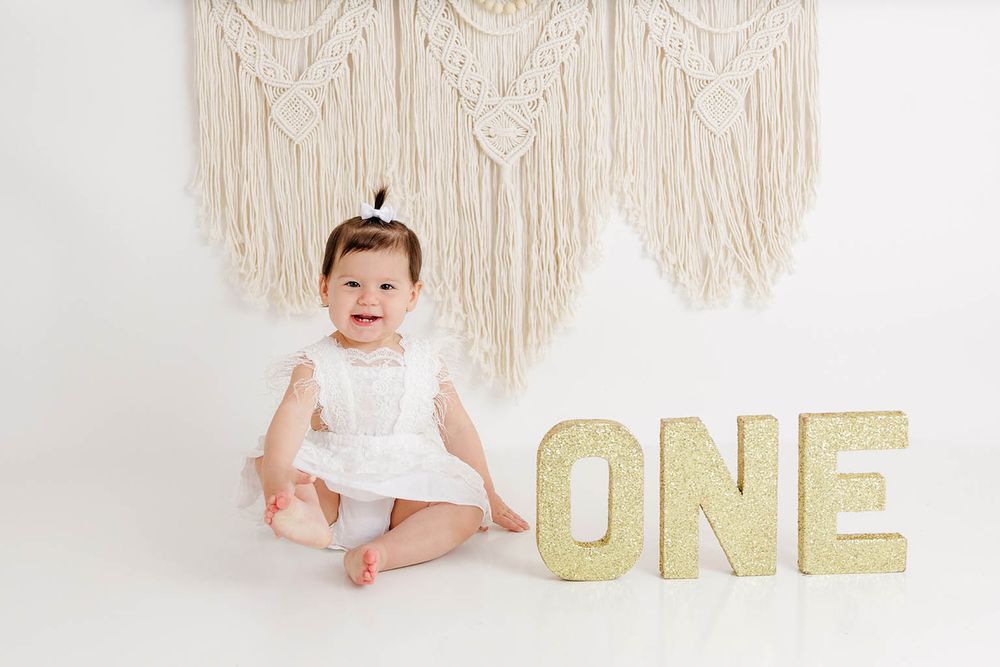 Happy baby girl in white dress laughing beside gold glitter ONE letters for first birthday portraits.