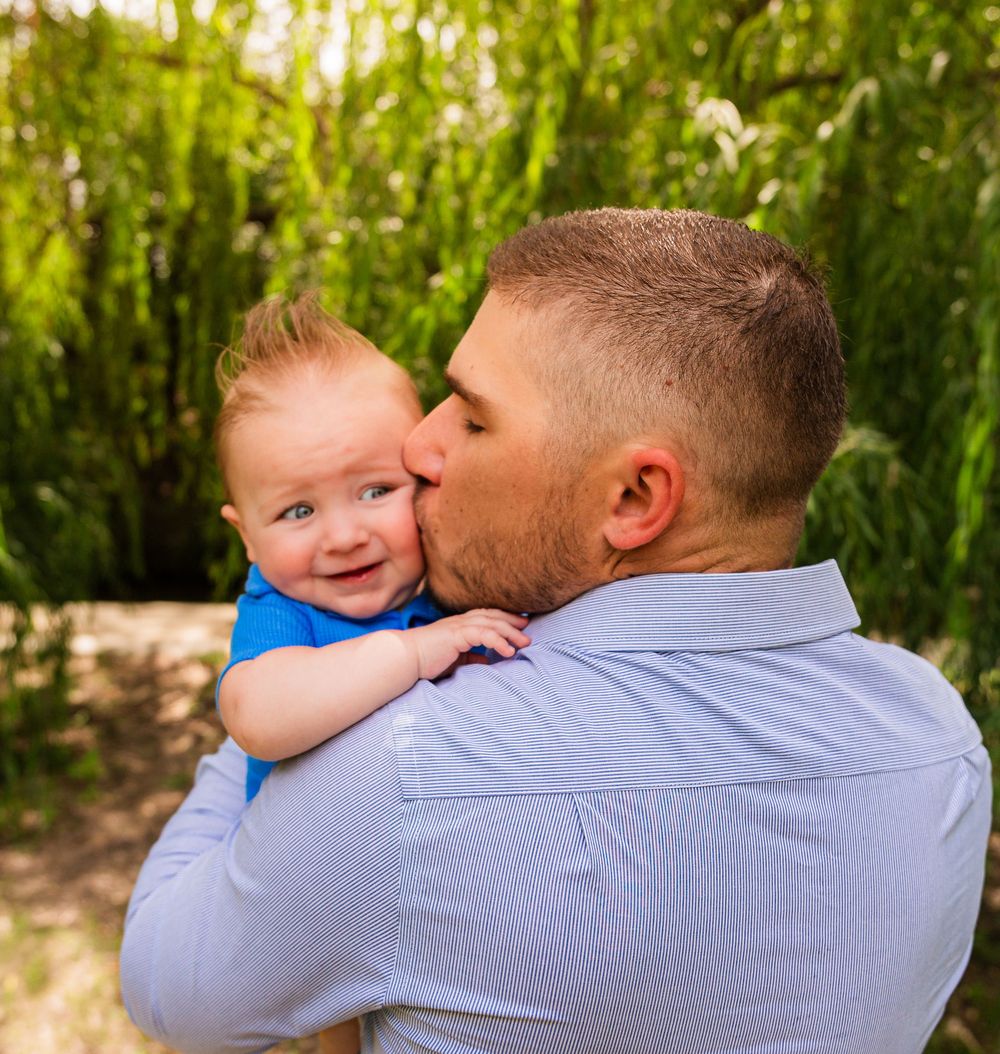 A man in a blue shirt kisses a smiling red-haired baby outdoors with lush green foliage in the background.
