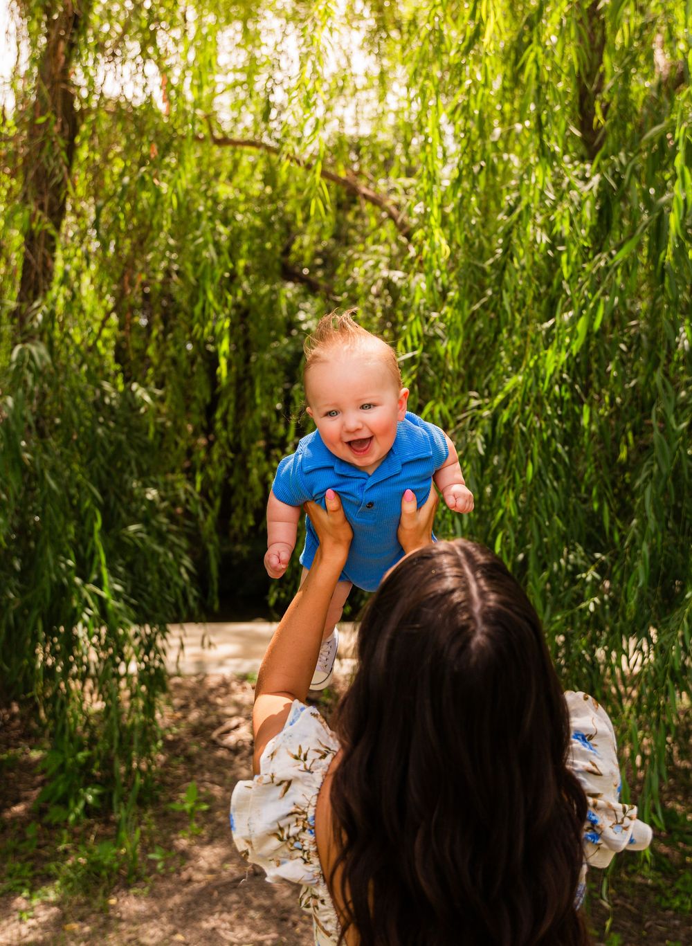 A joyful baby in a blue onesie being lifted by a dark-haired woman under lush green willow trees.