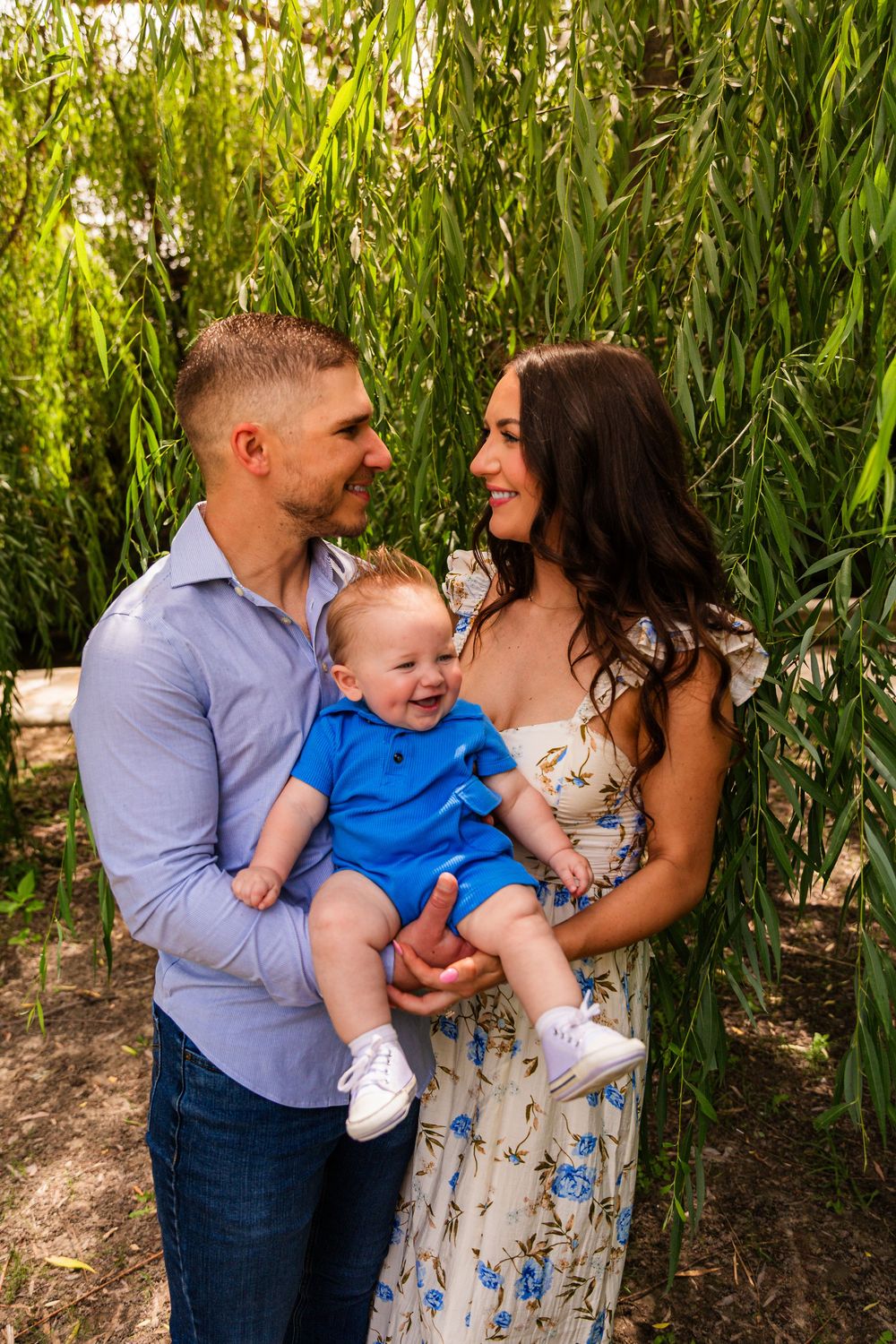 Happy young family with smiling baby in blue outfit posing under weeping willow tree outdoors.
