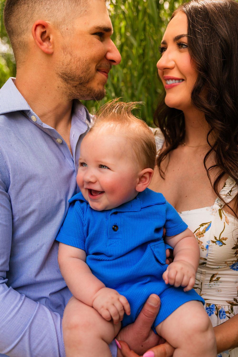 Happy family portrait with smiling baby in blue romper held by parents outdoors near green foliage.