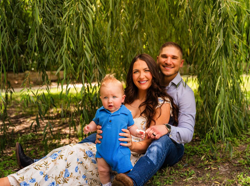 Young family with baby boy sitting under weeping willow tree, smiling outdoors in a lush green park setting.