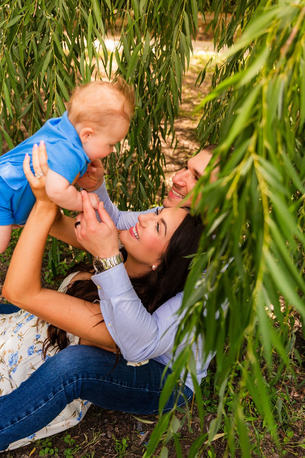 A smiling mother lifts her happy baby boy under a weeping willow tree outdoors.