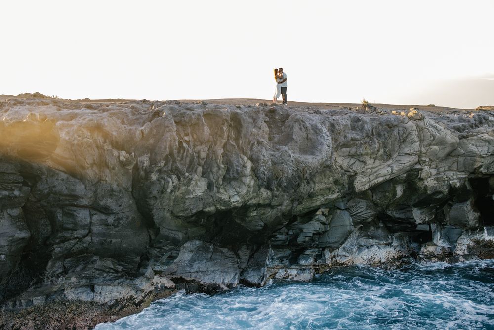 Couple embracing on rocky coastal cliffs at sunset with turquoise ocean waves crashing below.