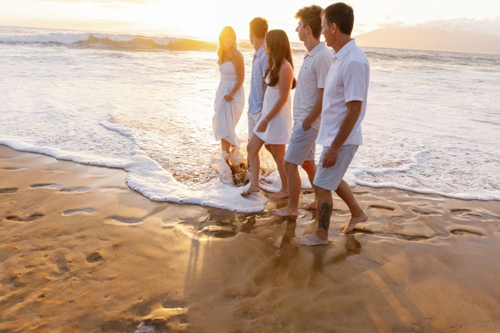 Four friends in white outfits walk barefoot along a sandy beach at golden sunset with ocean waves.