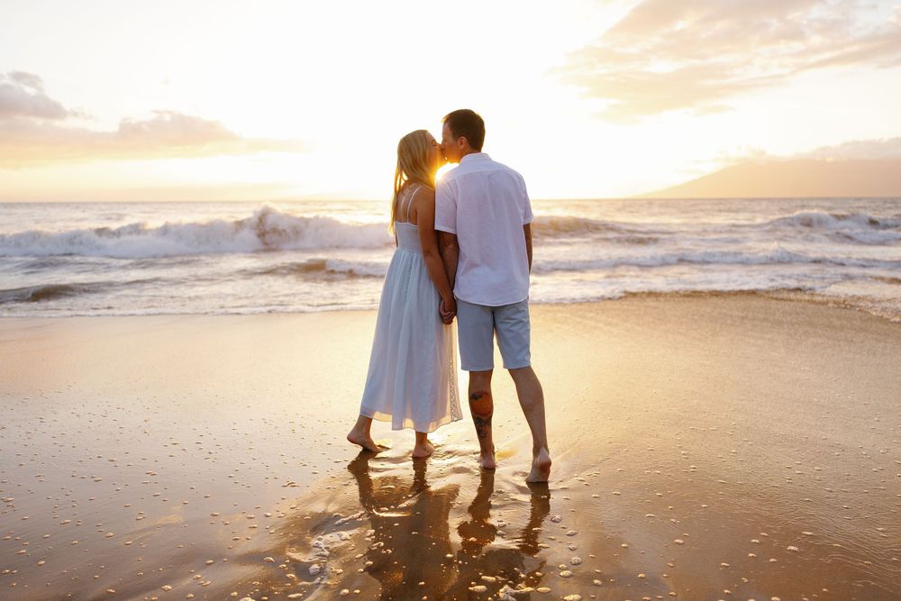 Romantic couple holding hands on a sandy beach at sunset, wearing white outfits with ocean waves behind them.