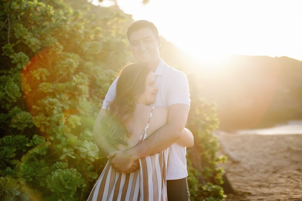 Couple embracing at golden hour on a sandy beach, bathed in warm sunset light with lush greenery behind them.