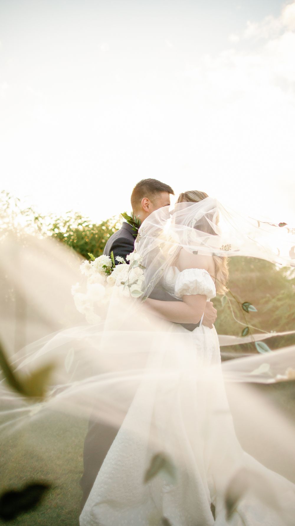 Bride and groom embrace outdoors in golden sunlight, with flowing white veil billowing in the foreground.