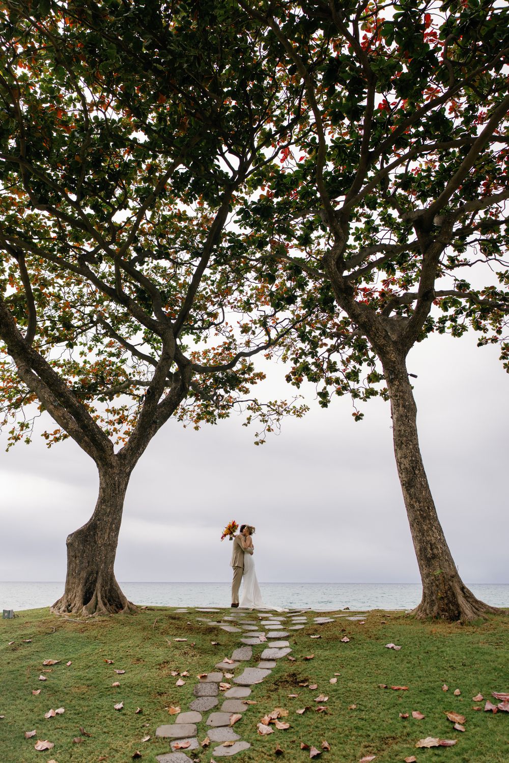 Bride and groom kissing between two large trees by the ocean on a cloudy day, with a stone path leading to them.