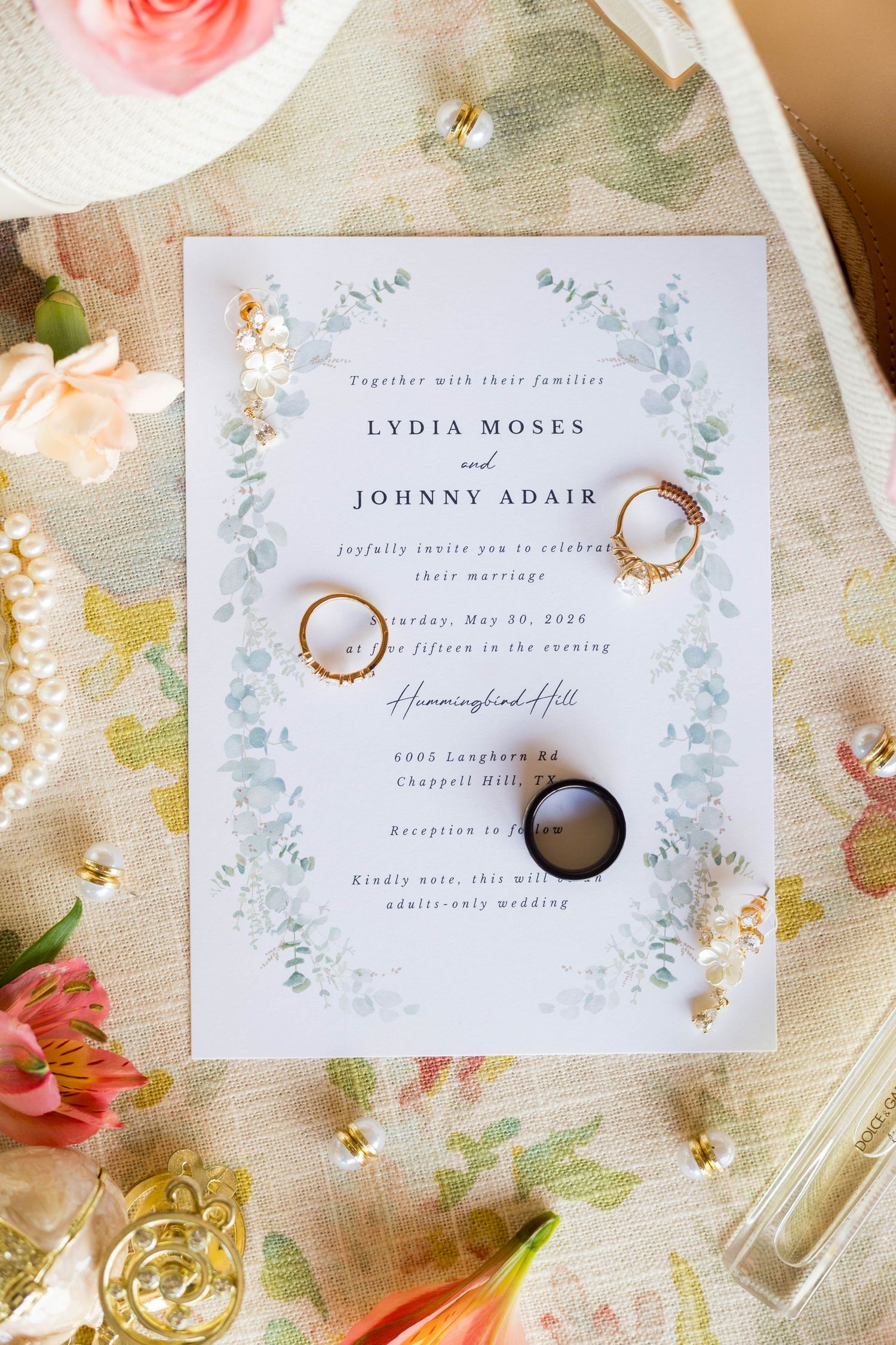 Wedding invitation for Lydia Moses and Johnny Adair at Hummingbird Hill, adorned with gold rings and floral decor.