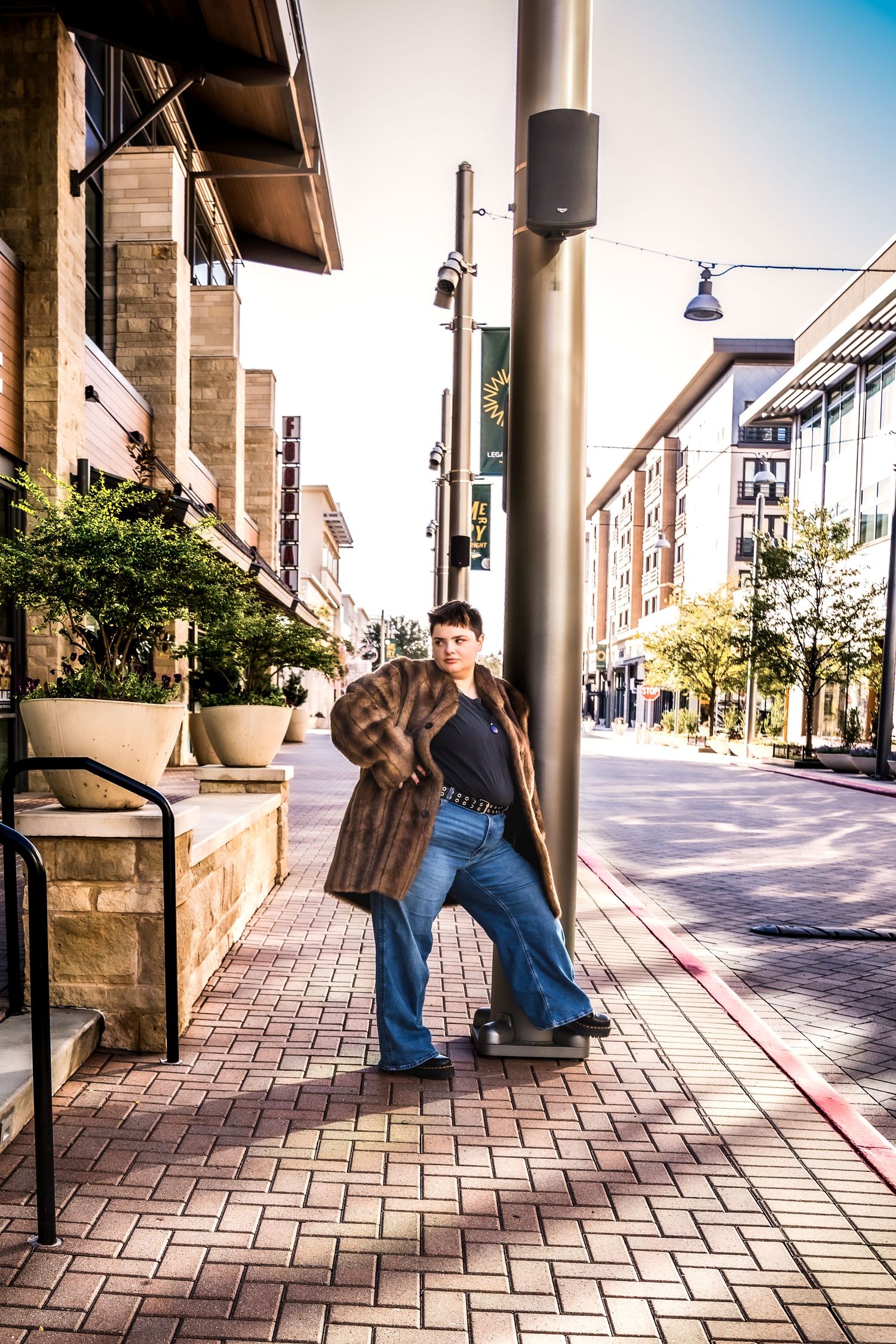 A young person in a fur coat and flared jeans leans against a pole on a sunny urban sidewalk.