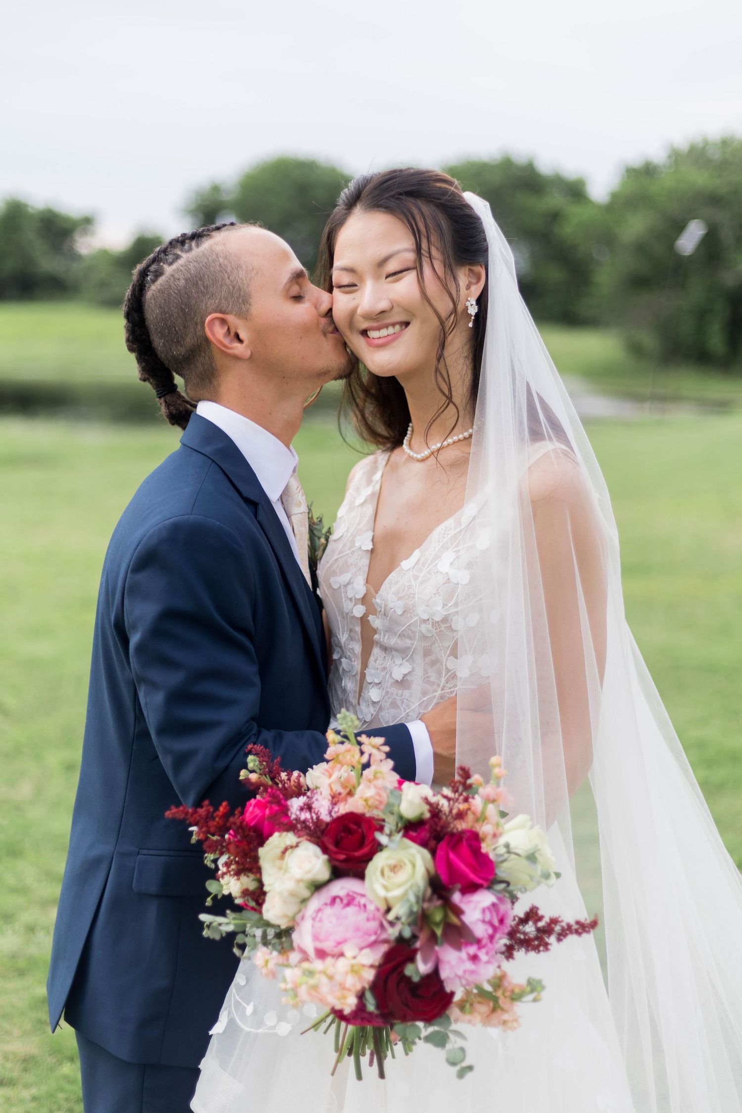 Groom in navy suit kissing bride in lace gown holding vibrant pink bouquet outdoors on wedding day.