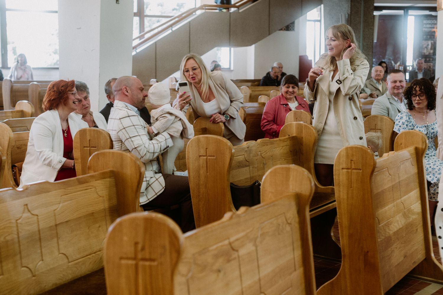 People socializing in wooden church pews with cross carvings during a casual gathering inside a bright church.