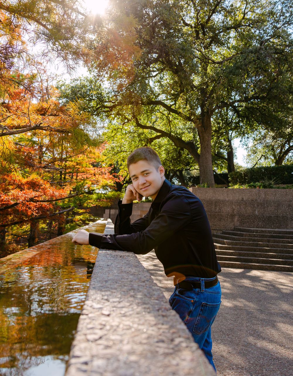 Young person leaning on stone ledge in autumn park, sunlight filtering through colorful fall trees.