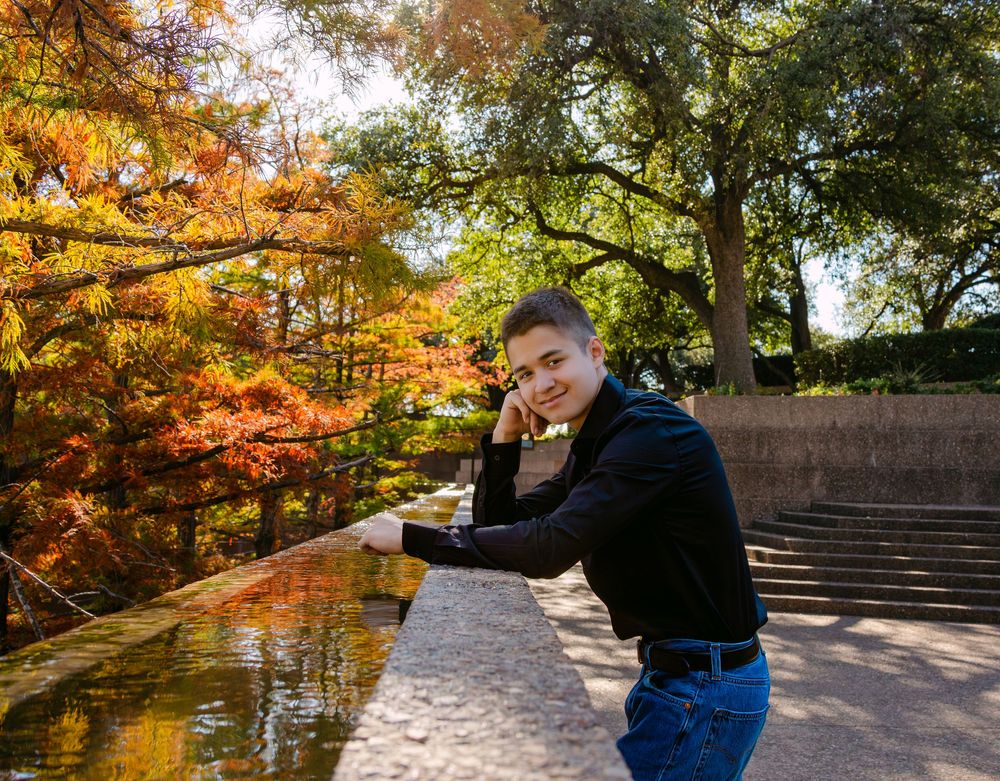 Young man leaning on stone railing in autumn park with colorful orange and yellow foliage trees.
