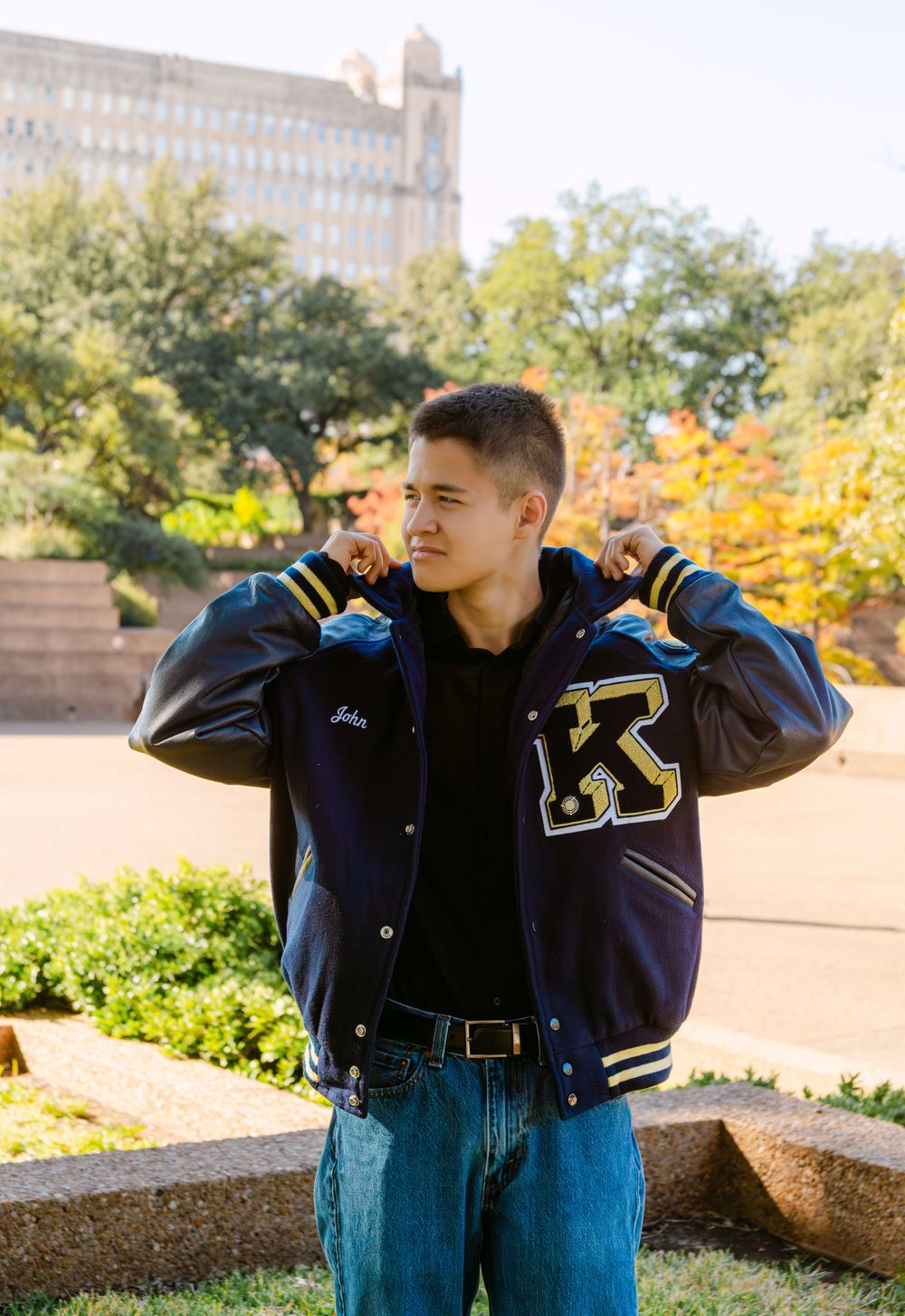 Young man wearing a navy blue varsity letterman jacket with K logo, posing outdoors near a building surrounded by autumn trees.