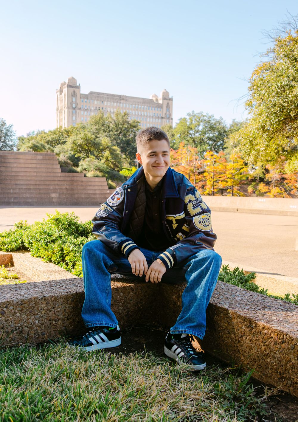 Young man in varsity jacket sitting on stone steps outdoors, smiling, with a historic building and trees in background.