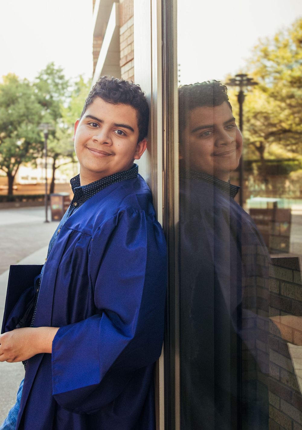 Young graduate in blue cap and gown smiling beside a reflective window outdoors.