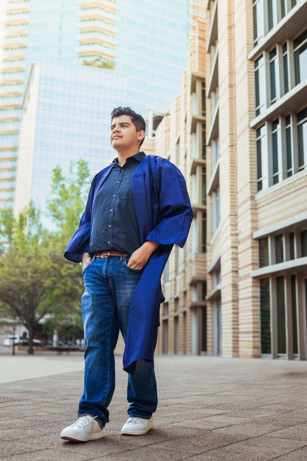 Young male graduate in blue academic gown stands confidently outdoors near modern buildings, looking upward with pride.
