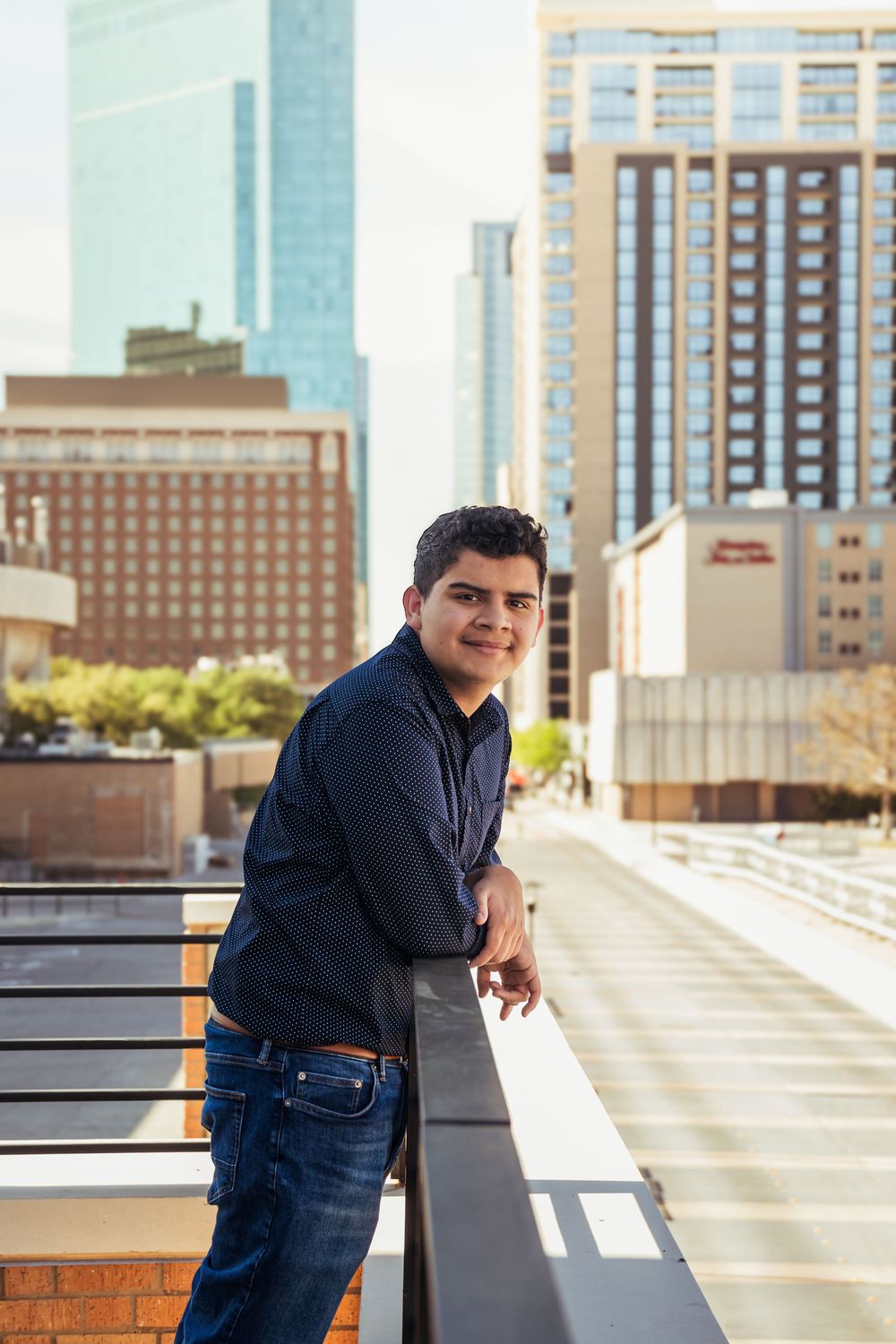 Young man in dark shirt and jeans leaning on railing with city skyline buildings in background.