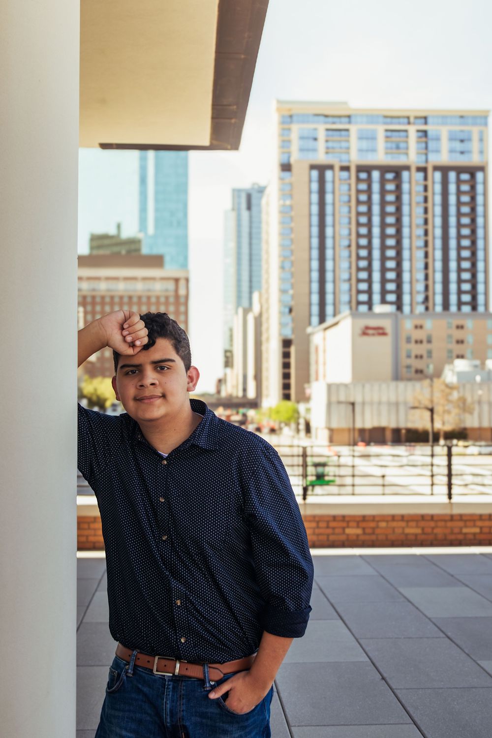 Young man in dark shirt leans against column with city skyline buildings visible in background.