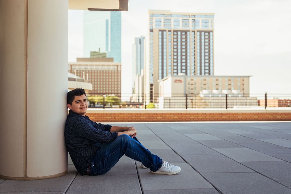 Young man sitting against a pillar on a rooftop terrace with city skyscrapers in the background.