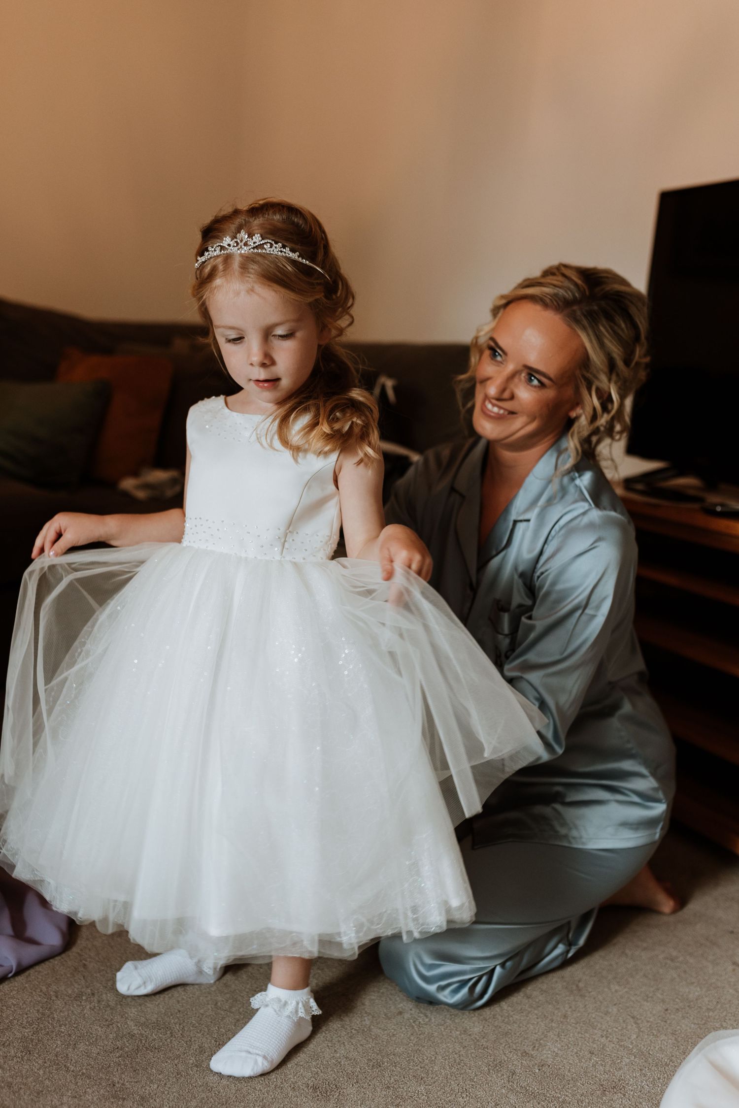 A woman in satin pajamas helps a young flower girl in a white tulle dress and tiara prepare for a wedding.