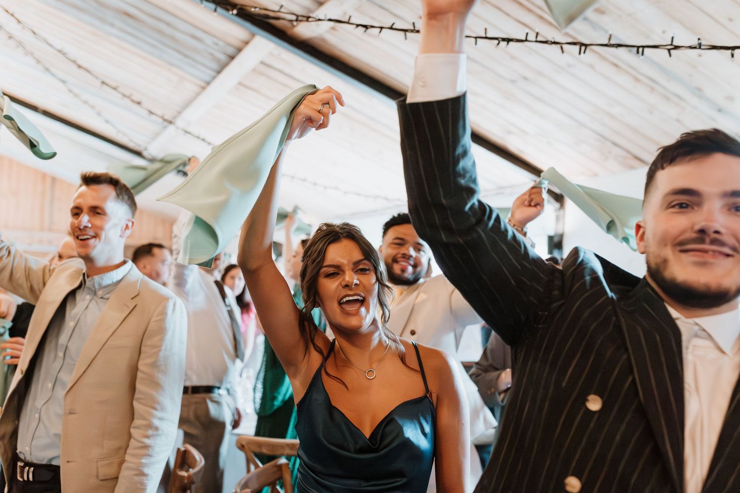 Guests celebrating at a wedding reception under a marquee, dancing and cheering with drinks raised.