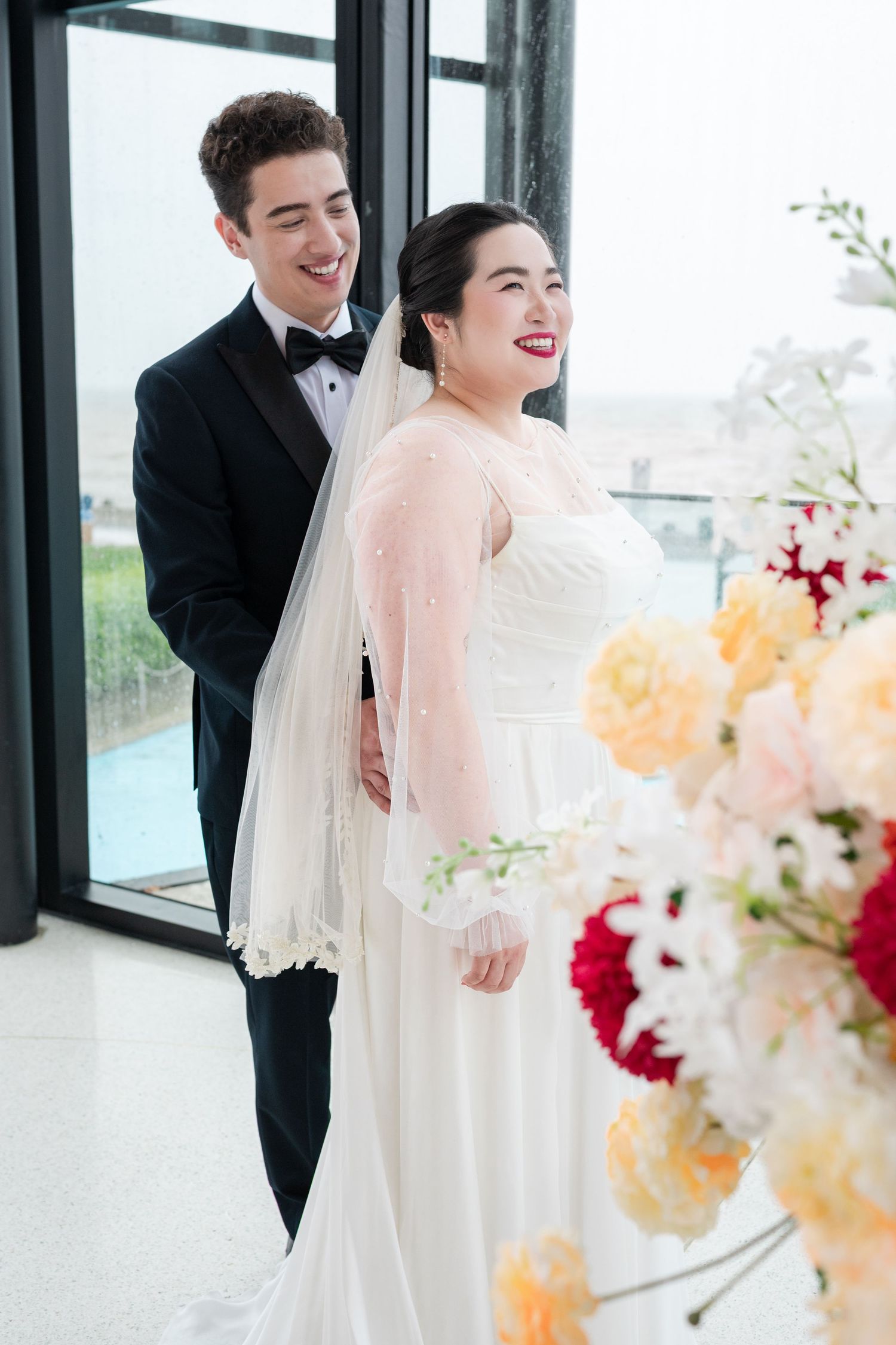 A smiling bride in a white gown and veil stands with a groom in a black tuxedo near floral arrangements.