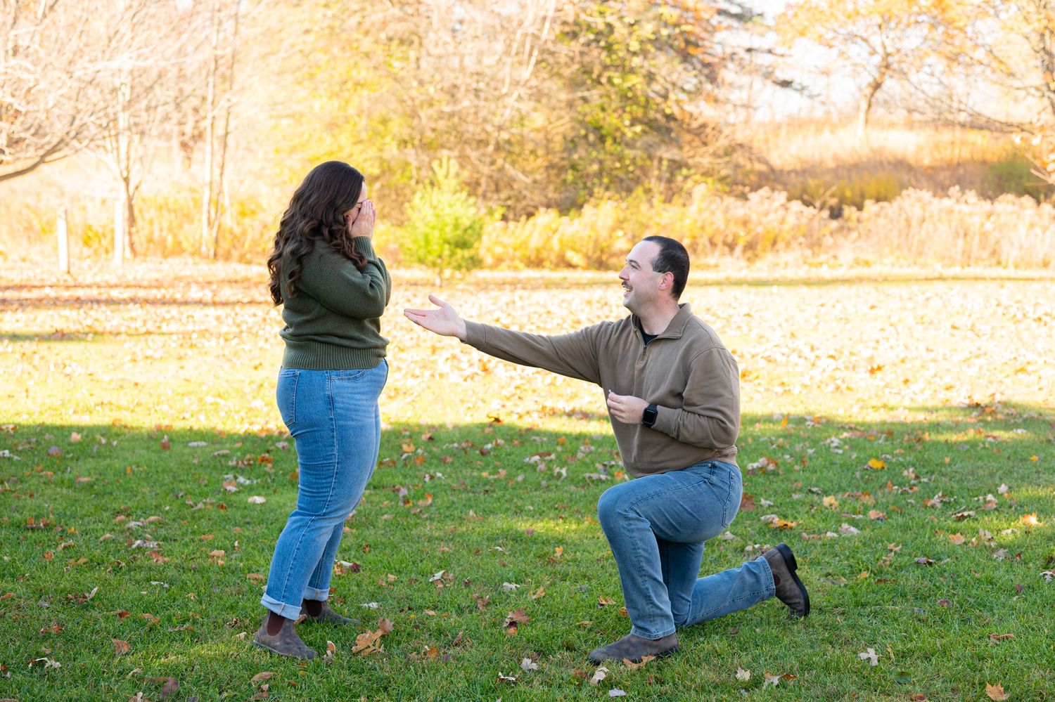 Man kneeling on one knee proposing to woman in sunny autumn park with colorful fall foliage backdrop.