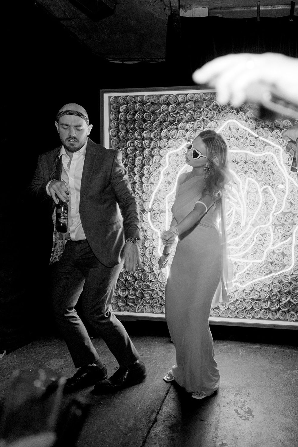 Black and white photo of a couple dancing at a wedding reception in front of a floral neon rose backdrop.