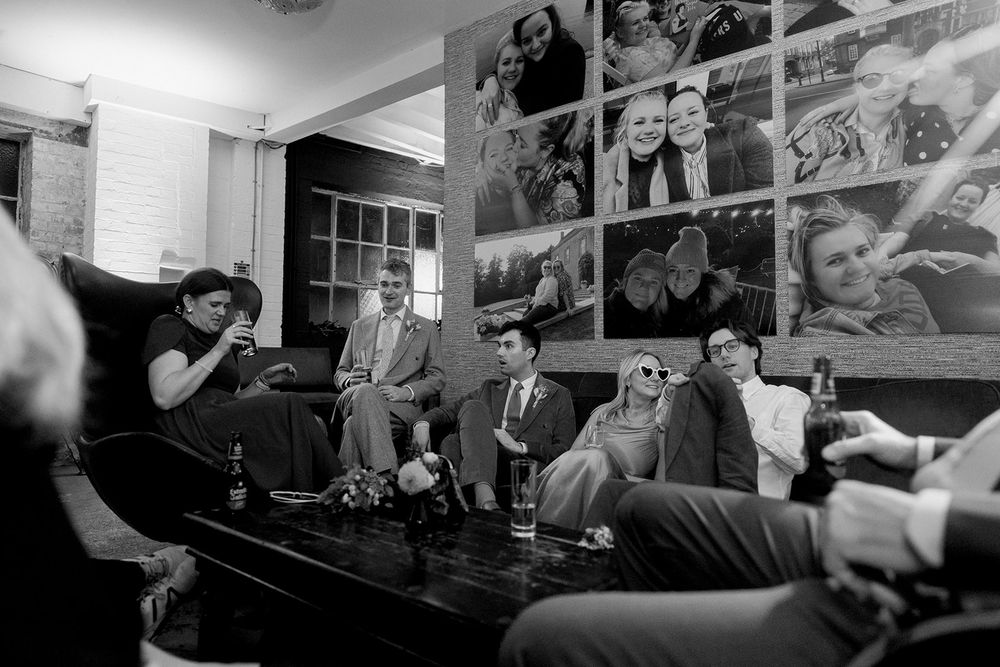 Black and white photo of wedding guests relaxing on sofas with photo wall backdrop at indoor reception.
