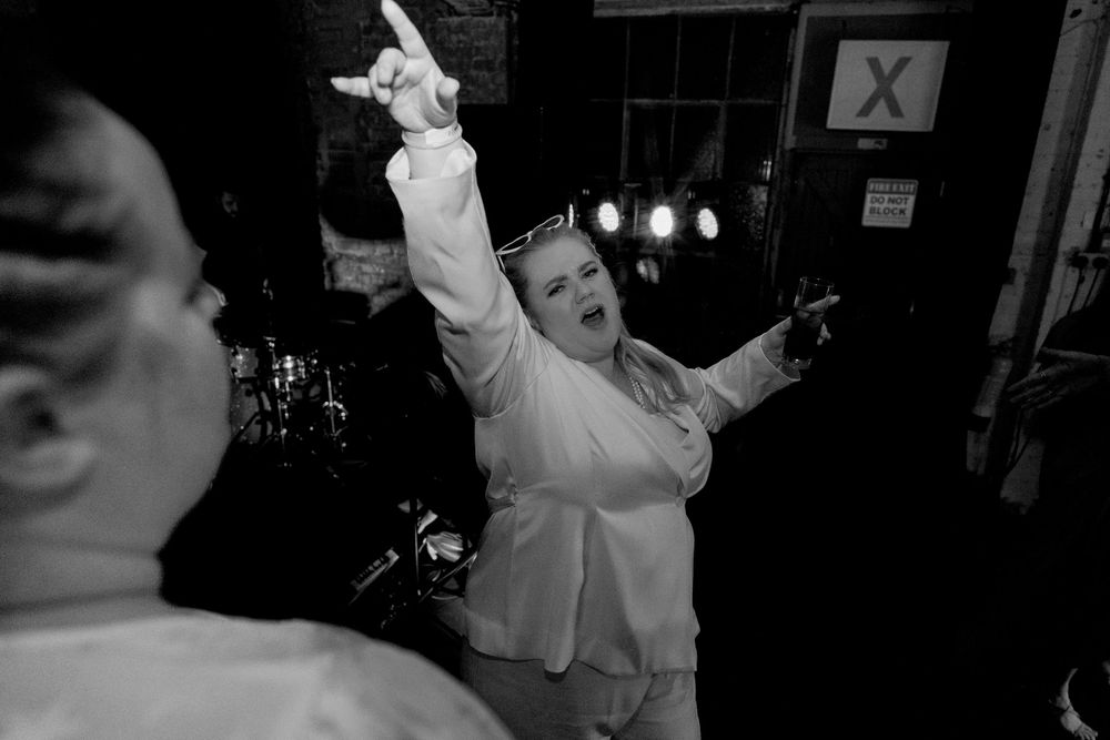 Black and white photo of a woman in white outfit dancing energetically with arm raised at a lively wedding reception.