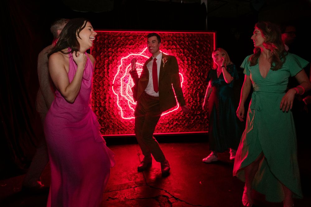 Guests dancing joyfully at a wedding reception with a glowing red neon sign backdrop in a dark venue.