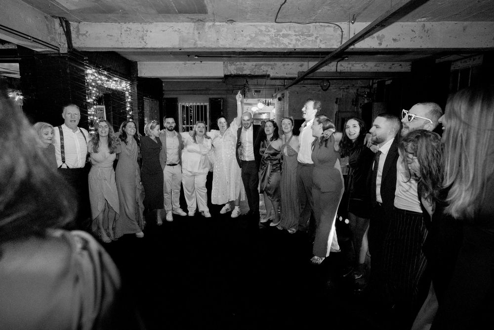 Black and white photo of a large group of elegantly dressed guests gathered on a dance floor at an indoor wedding reception.