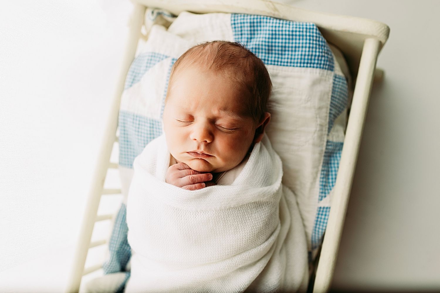 Peaceful newborn baby swaddled in white blanket sleeping in a small wooden crib with blue checkered bedding.