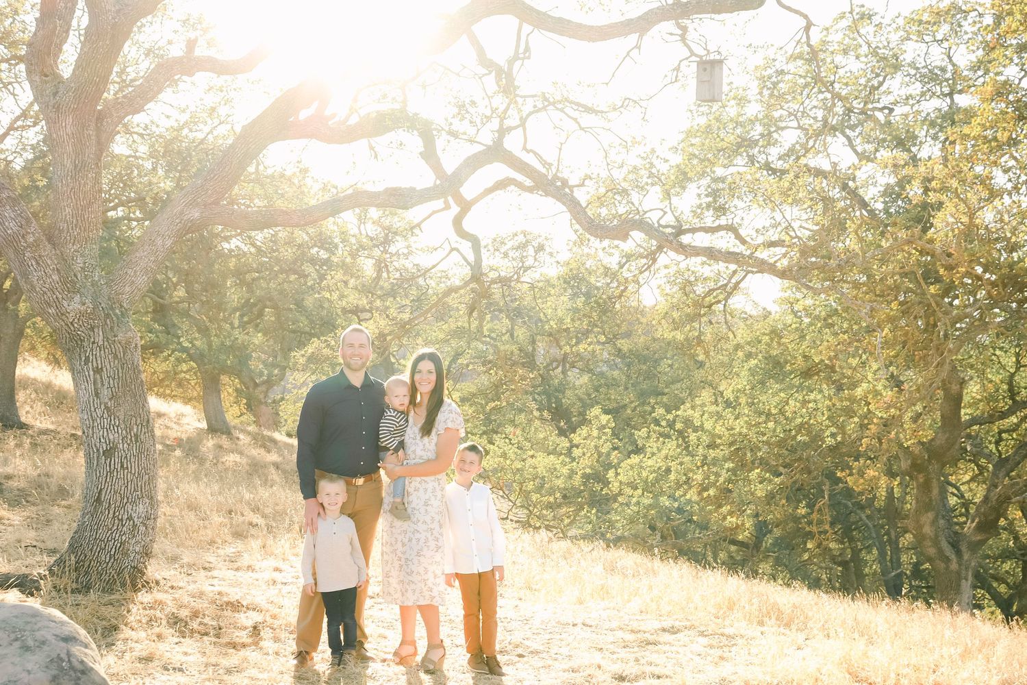 Family of five posing together outdoors among oak trees in golden sunlight on a dirt trail.