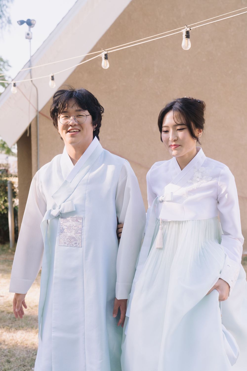 Couple wearing traditional white Korean hanbok attire standing together outdoors at a wedding ceremony.