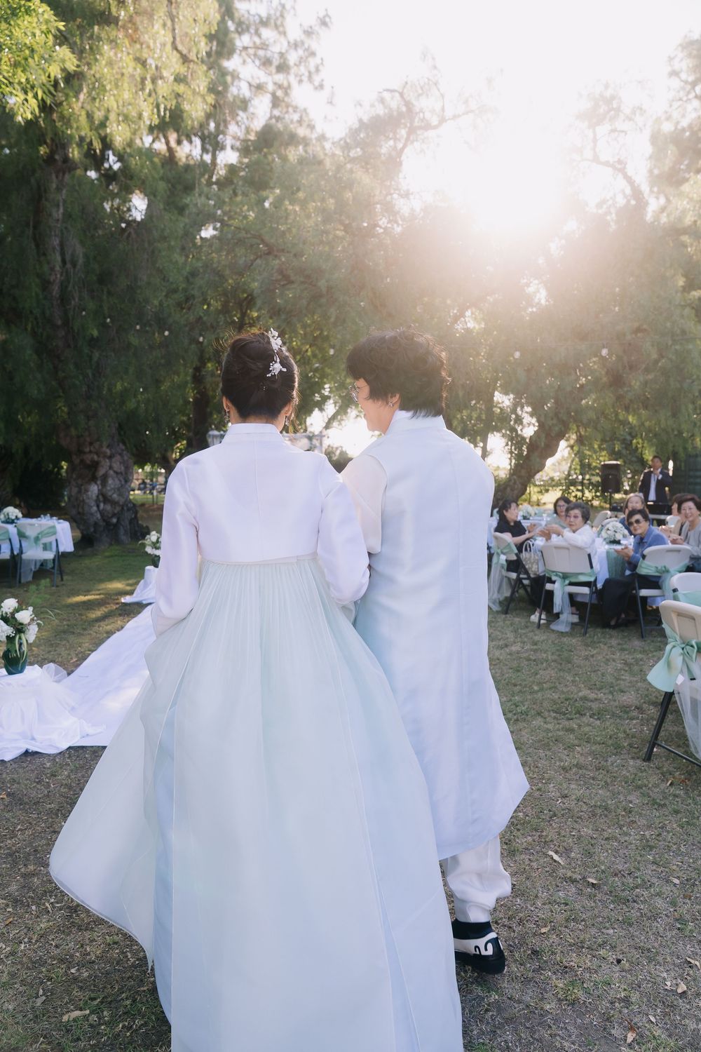 Couple in traditional Korean hanbok wedding attire walking toward outdoor reception under sunlit trees.