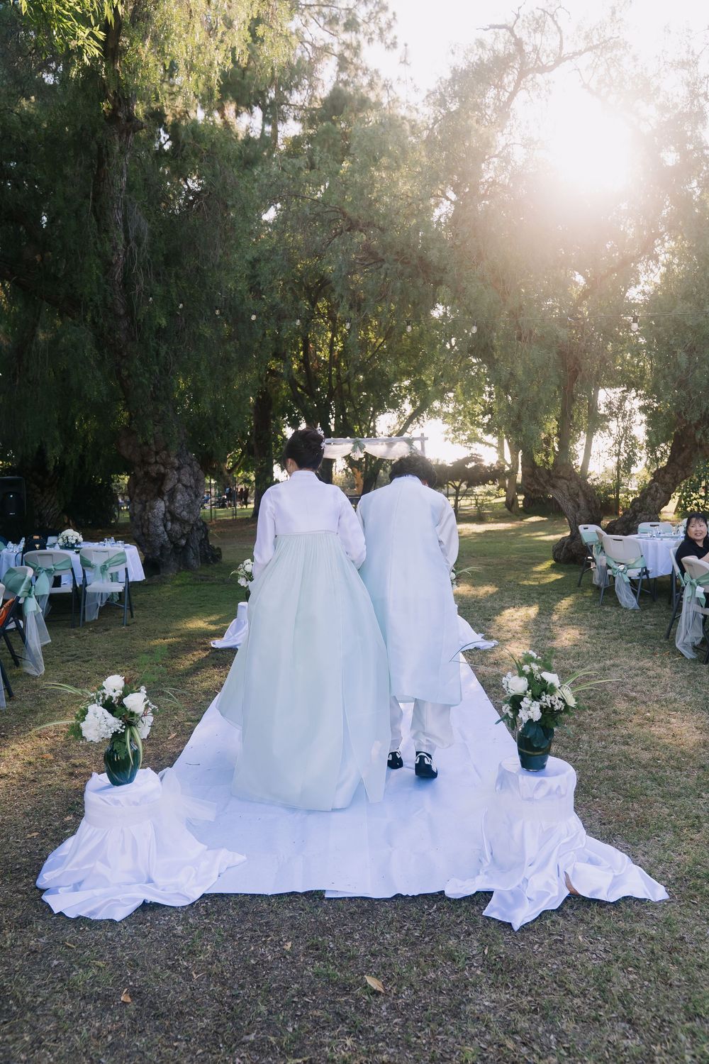 Bride and groom in traditional Korean hanbok walking toward sunlit outdoor wedding altar surrounded by trees.