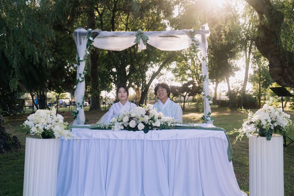 Two people sit at a white-draped wedding table adorned with white roses under a floral canopy outdoors.